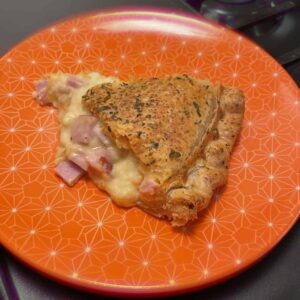 A single slice of macaroni and cheese pie sits in the middle of a decorative orange place with a white geodesic pattern on it. The pie has a nicely seasoned flaky crust and you can see the melted cheese, noodles and ham gently flowing onto the plate.