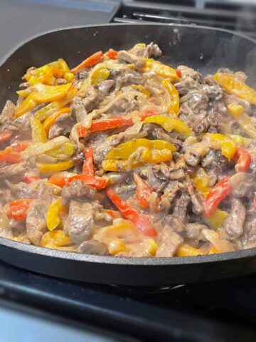 View of a skillet cooking sliced steak, peppers and onions in a cheesy sauce.